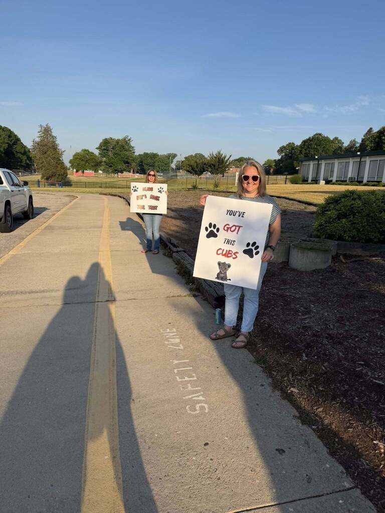 Two women stand outdoors on a paved area holding supportive signs for students. The woman on the right holds a sign reading "YOU'VE GOT THIS CUBS" featuring paw prints and a drawing of a lion cub. The woman on the left holds a sign reading "CUBS LET'S ROCK THE TEST" with paw prints. On the sidewalk in front of them, the words "SAFETY ZONE" are written vertically in chalk. The scene is set outside a school with a road/driveway, grass, trees, and buildings visible in the background under a clear sky. Long shadows suggest early morning or late afternoon lighting. A white truck is parked on the left.