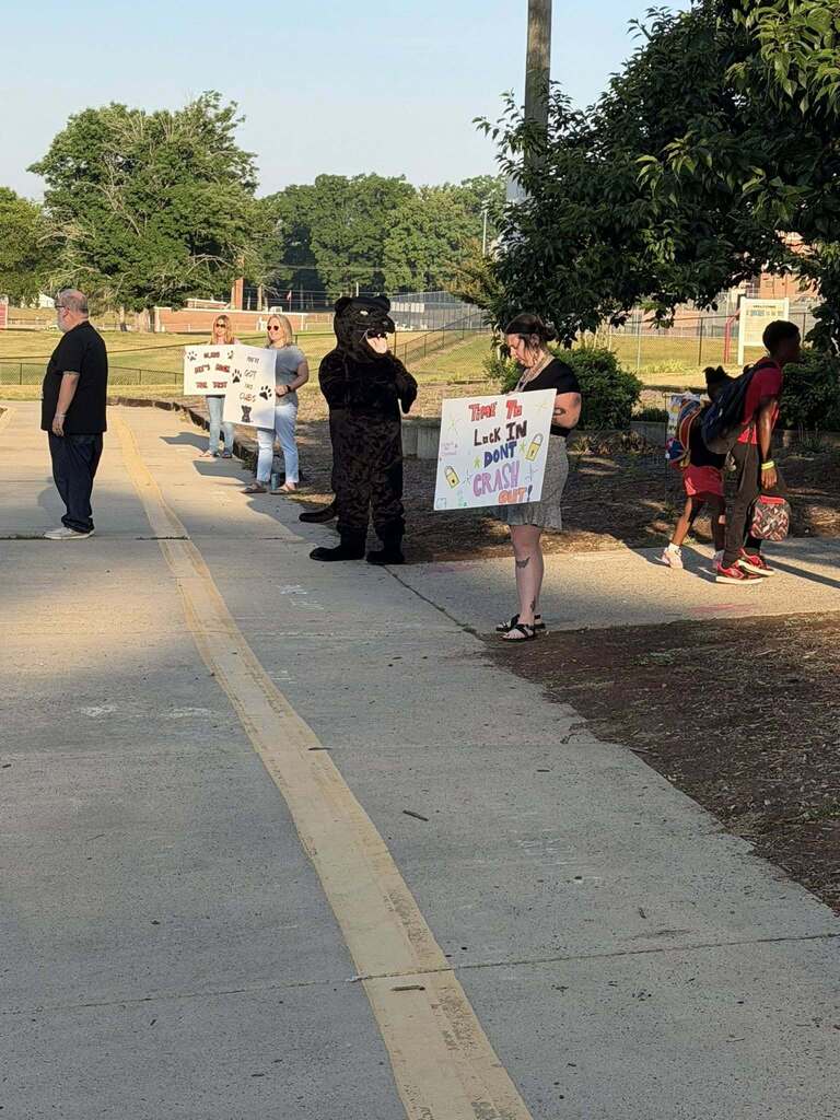 A group of five adults and two children gather on a paved path outdoors near a school. One adult is dressed in a black panther mascot costume. Two women hold signs with paw print graphics and text supporting "cubs," while another woman holds a sign reading "TIME TO LOCK IN & DON'T CRASH OUT!" with padlock and star graphics. A man stands on the far left, and two children with backpacks walk past the group on the right. The signs collectively display messages "WE LOVE OUR CUBS!" and "WE'VE GOT THE BEST CUBS!" Trees and a fence are visible in the background.