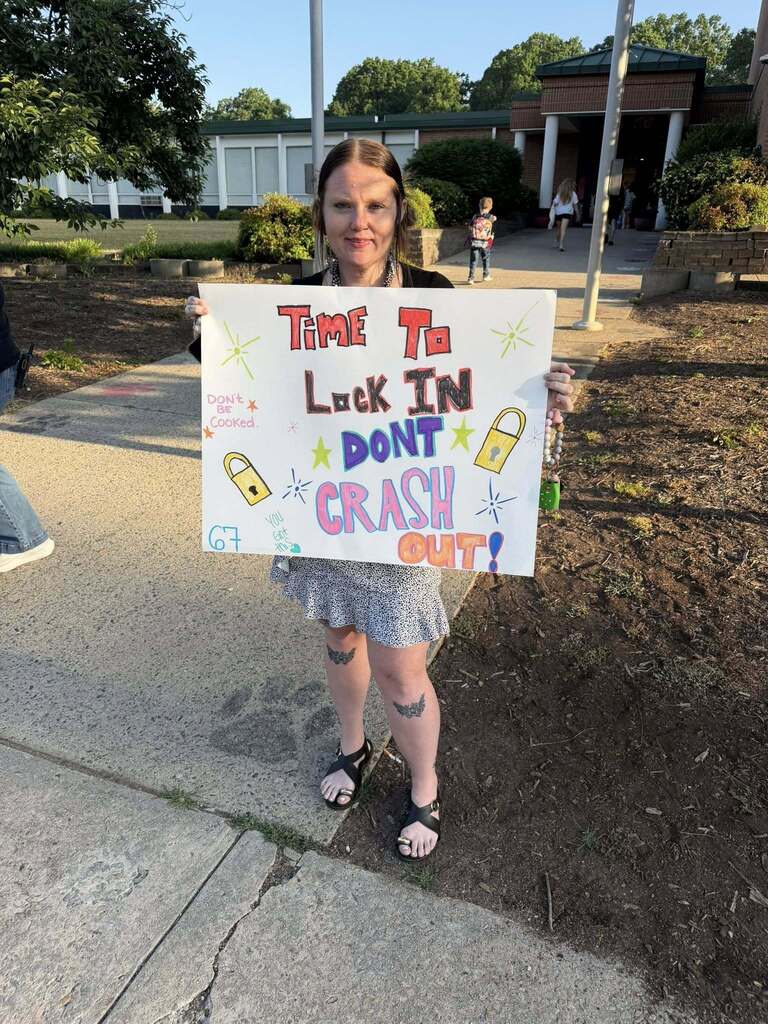A woman stands outdoors holding a white poster board sign with colorful, hand-drawn text and graphics. She has long dark hair and wears a black top, a patterned skirt, and black sandals, with bat-wing tattoos visible on her knees. The sign reads "TIME To Lock IN DONT CRASH OUT! DON'T BE Cooked. 67 You Got This" with decorative drawings. In the background, a school building is visible with trees and a sidewalk. A child with a backpack and another person are visible in the distance near the building's entrance. The bright, clear sky indicates daytime.