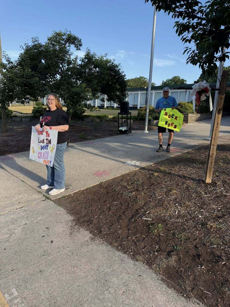 A woman and a man stand on a paved walkway outdoors holding supportive signs for an EOG (End of Grade) testing campaign. The woman on the left smiles while holding a white sign with colorful text and lock drawings reading "Time To Lock IN DON'T CRASH OUT!" The man on the right holds a yellow sign with black and red text and musical notes