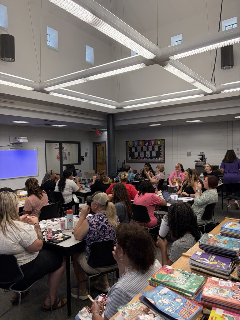 A large group of people are seated at tables in a modern classroom or educational facility, facing toward the front of the room for what appears to be a group event or meeting. The space features contemporary design elements including a ceiling with integrated linear LED lighting fixtures arranged in a grid pattern and multiple small windows in the upper walls providing natural light. Room Features: A large display screen is mounted on the left wall showing blue content. On the right wall, a bulletin board displays what appears to be a chess-themed exhibit. Stacks of books are visible in the foreground on the right side. The room has white walls and is well-lit with modern lighting. Participants wear casual clothing, and the setting suggests a school or educational facility hosting a professional development session or staff meeting.