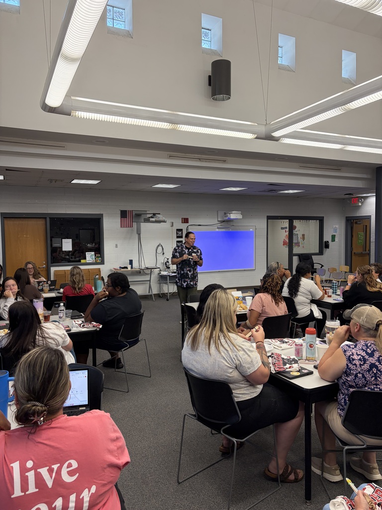 A professional development or educational workshop takes place in a modern institutional classroom or training space. Approximately 20-25 attendees are seated at multiple round tables throughout the room, eating and participating in the session. A man in a floral patterned shirt stands near a wall-mounted interactive display screen showing blue content, appearing to facilitate the presentation or discussion. Room Features: The space has a high ceiling with LED linear lighting, skylights, and a black pendant light fixture. White walls and gray concrete flooring create a contemporary atmosphere. Large windows provide natural light. An American flag and various framed educational materials are displayed on the walls. Visible infrastructure includes air ducts, electrical outlets, and exit signage. The modern educational setting appears designed for collaborative workshops, training sessions, or team-building events.