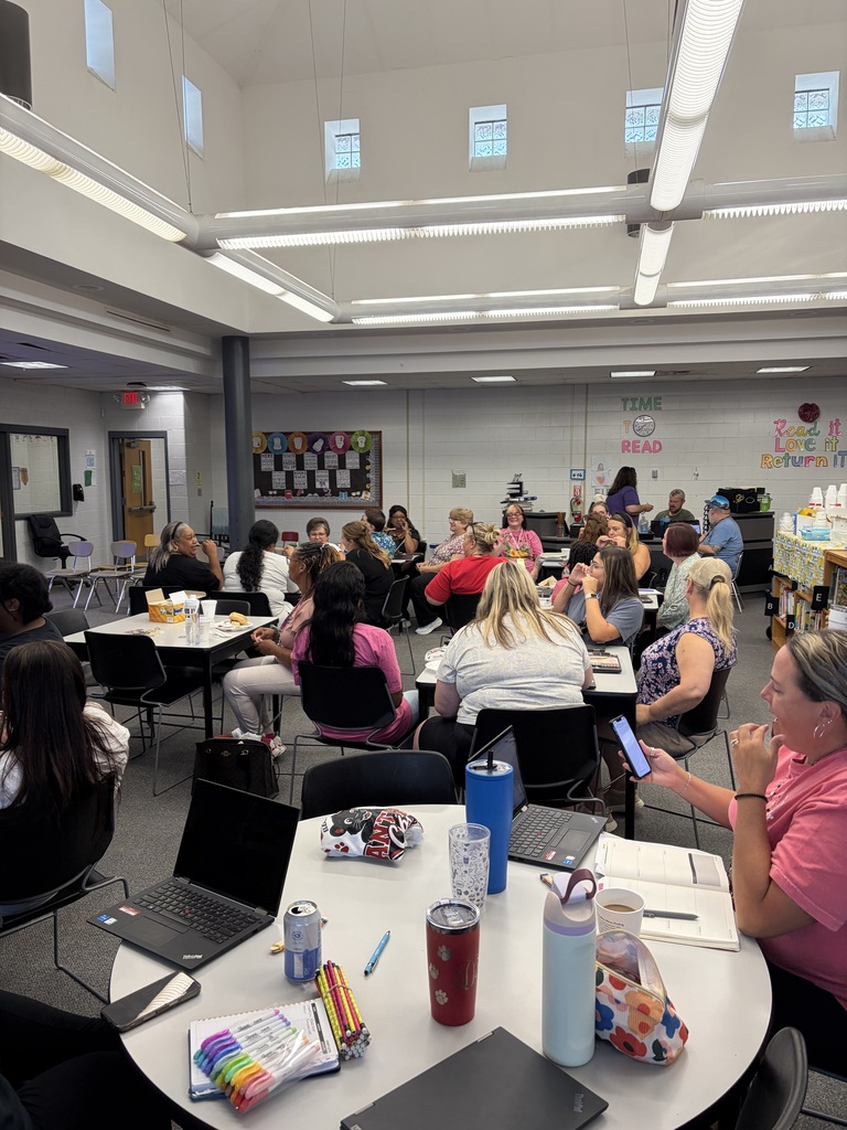 Approximately 30-40 educators or staff members are seated at tables in a school library or media center for what appears to be a professional development meeting, workshop, or collaborative planning session. Tables are equipped with laptops, notebooks, writing supplies including visible colored markers, and beverages, indicating an active working session. Room Features: The bright, well-lit space features modern fluorescent ceiling lighting and large windows along the upper walls. Educational displays are visible throughout the room, including a bulletin board and reading-themed signage displaying "TIME TO READ" and "Return it." Contemporary classroom furniture and organizational systems are visible in the background. The setting creates a collaborative professional learning environment typical of educator training or planning sessions.