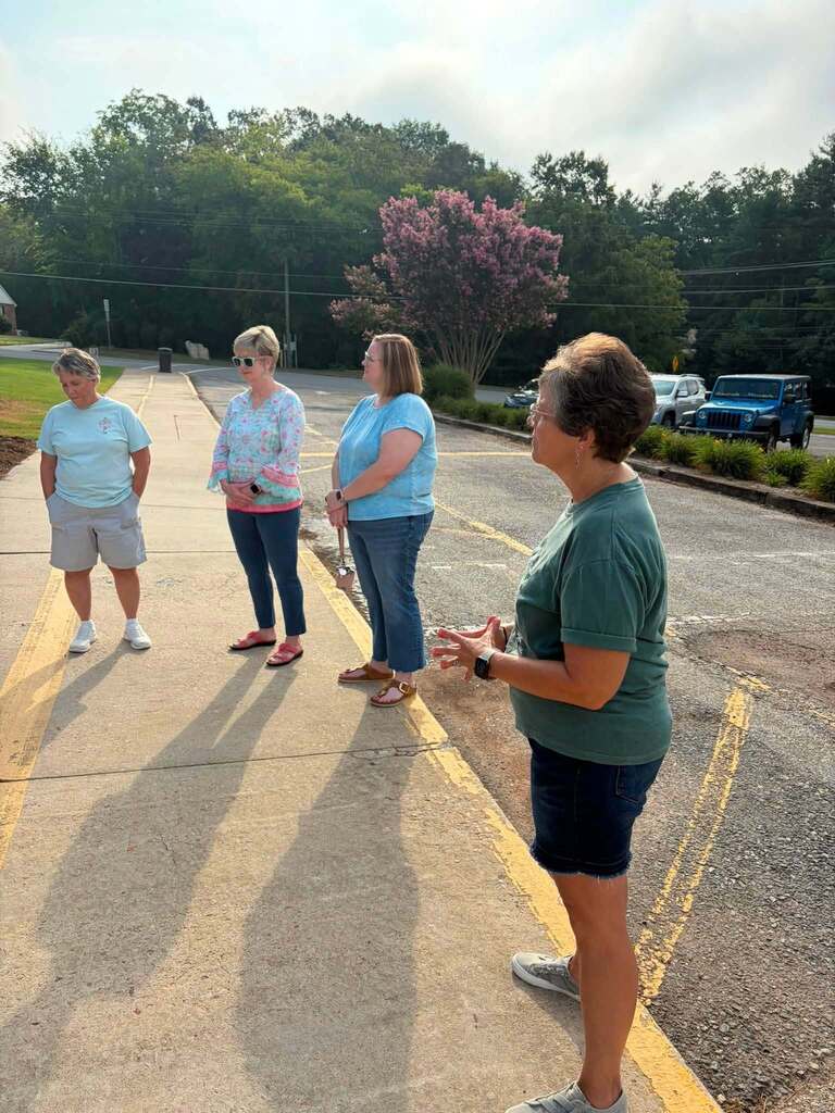 Four women gather in a parking lot during daytime for what appears to be a casual outdoor meeting or group activity. A woman in a green shirt on the right appears to be speaking or leading a discussion with the other three women, who wear light blue and patterned shirts. They stand on a paved surface marked with yellow lines. The background features a pink-flowering tree, green vegetation, parked cars including a blue Jeep, utility poles, and overhead power lines under a partly cloudy sky.