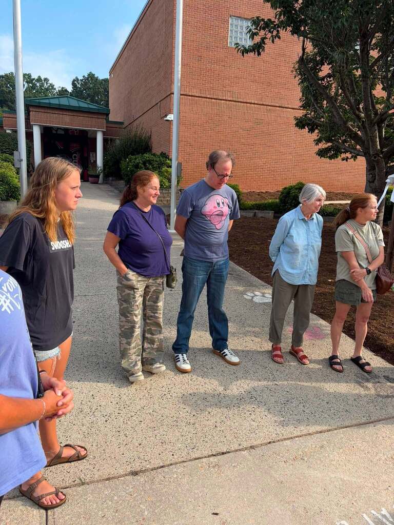Five people of various ages—from teenagers to seniors—stand outdoors in front of a large red brick building for what appears to be a tour or outdoor meeting. The building features a covered entrance visible to the left, suggesting an institutional or church setting. Mature trees and landscaping surround the area under a clear blue sky. The group, dressed casually in t-shirts and pants, stands on a paved concrete surface engaged in conversation or listening to a presentation.