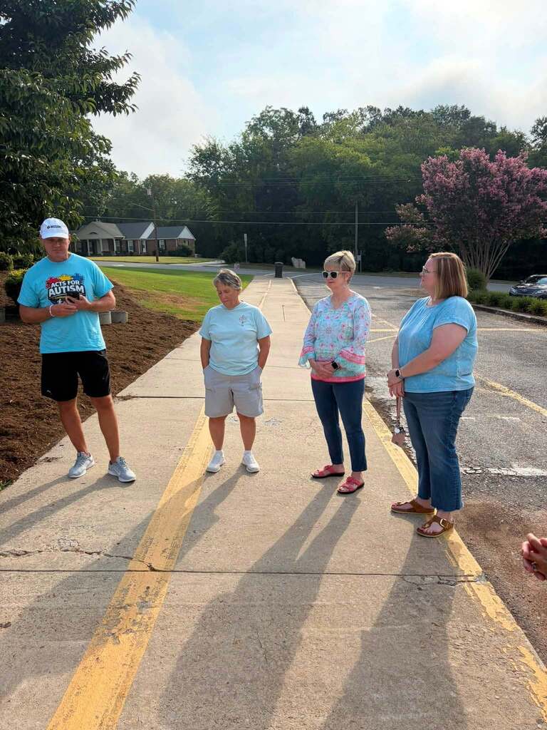 Four people stand on a parking lot with yellow road markings, participating in what appears to be a charity or community event. The man on the left wears a blue t-shirt reading "ACTS FOR AUTISM," indicating the event's charitable purpose. The group is positioned on a lined parking area in front of a brick building. The background features pink flowering trees, green trees, and flowering shrubs creating a peaceful, suburban setting with manicured landscaping. The sky is clear with scattered clouds.