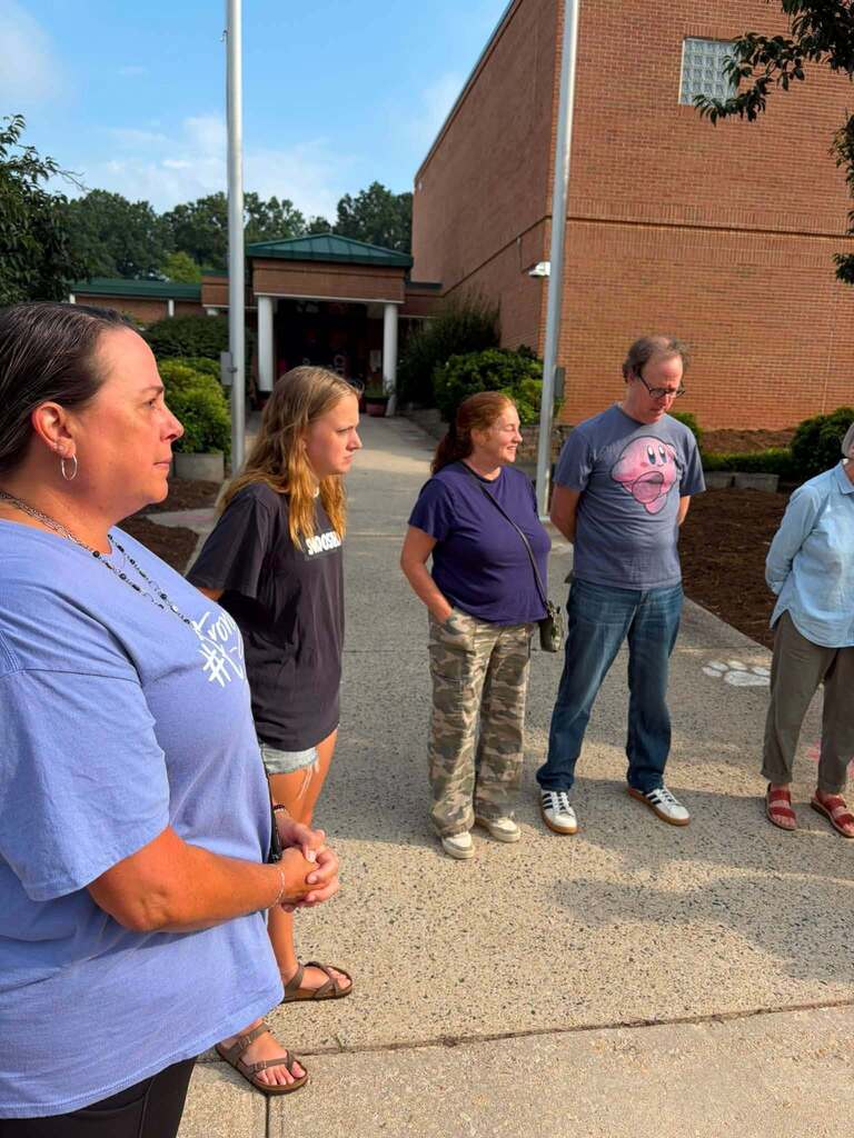 Five people stand outdoors on a paved walkway in front of a brick institutional or educational facility building with a covered entrance visible in the background. The group gathers in a casual formation, appearing to listen to or engage with a woman on the left wearing a light blue t-shirt with orange accents. Other group members wear casual clothing including dark shirts and patterned pants. Trees and landscaping are visible in the background under a mostly clear blue sky. The setting suggests a tour, meeting, or outdoor educational gathering taking place.