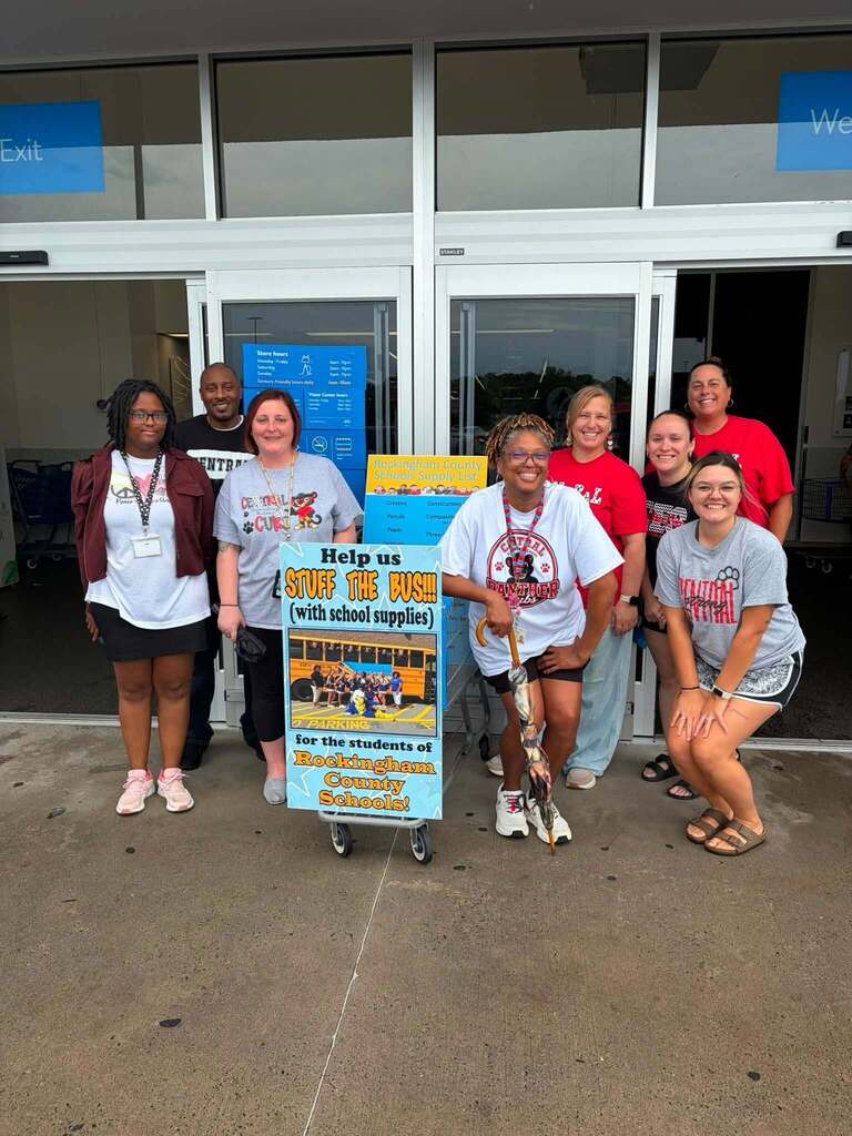 7 people stand together outside a modern building with large windows and white trim, posing in front of a decorated promotional sign mounted on a wheeled cart. The sign reads "Help us STUFF THE BUS (with school supplies)" and features an image of a school bus, with text indicating the initiative is "for the students of Rockingham County Schools!" The group is smiling and appears to be staff or volunteers participating in a school supply drive. The setting is outside a community building or school facility, with "Welcome" text visible on the upper right window.