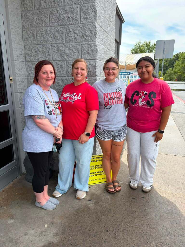 Four young women stand together outdoors against a gray concrete block building, posing for the camera and smiling. They wear casual clothing including t-shirts in light blue, red, gray, and pink/magenta colors. The setting appears to be outside a commercial or institutional building with a parking area visible in the background and trees visible in the distance. The sky is partly cloudy. A yellow warning sign is partially visible on the ground near them.