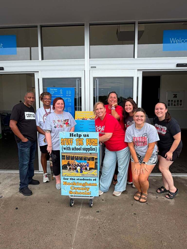 Eight people pose together outside a building with glass doors and windows, participating in a school supply drive for Rockingham County Schools. They stand in front of a large promotional sign on a wheeled cart reading "Help us STUFF THE BUS (with school supplies) for the students of Rockingham County Schools!" in bold yellow and blue lettering, featuring an image of a school bus below the text. The group, appearing to be staff or volunteers, is dressed casually in t-shirts and smiling at the camera. The setting is outside a store or community center entrance, with "Exit" signage visible on the storefront windows.