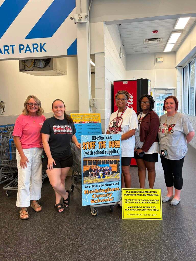 Five people stand together in an indoor facility posing with a collection cart and promotional signage for a school supply drive. The space features fluorescent lighting, white walls, industrial-style ceiling fixtures, and "CART PARK" signage with blue and white stripes visible on the wall.