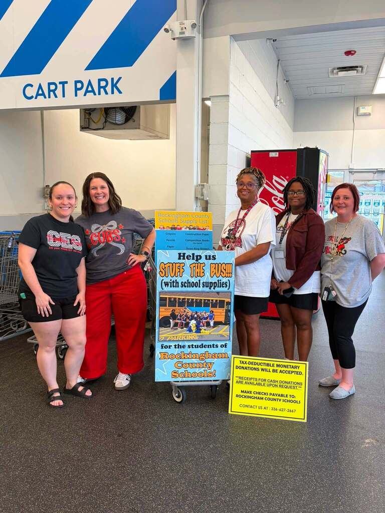 Five people stand in a cart park area (indicated by a "CART PARK" sign with blue and white diagonal stripes visible above) next to a promotional display for a school supply donation drive. The main blue and yellow poster reads "Rockingham County Schools Buying List Presents Community Books - Help us STUFF THE BUSH! (with school supplies) for the students of Rockingham County Schools!" A yellow placard below states "TAX-DEDUCTIBLE MONETARY DONATIONS WILL BE ACCEPTED. 'RECEIPTS FOR CASH DONATIONS ARE AVAILABLE UPON REQUEST.' MAKE CHECK PAYABLE TO: ROCKINGHAM COUNTY SCHOOLS! CONTACT US AT: 336-627-2647."