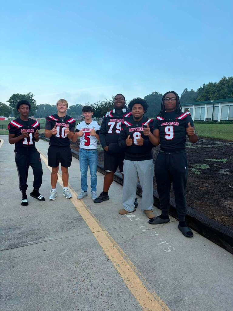Six young people stand together on a parking lot or road giving thumbs-up gestures. Most wear black Panthers sports uniforms with red and white stripe trim, with visible jersey numbers including 11, 10, 5, 79, 8 and 9. One person wears a white Panthers jersey. The background features trees, a clear blue sky, and a white building structure, suggesting the setting is a school or sports facility.