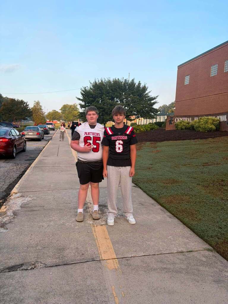 Two teenage boys stand on a concrete sidewalk with a yellow center line in front of a school building. The boy on the left wears a white and red "Panthers" jersey numbered 65. The boy on the right wears a black and red "Panthers" jersey numbered 6. The setting features a large red brick school building with windows on the right, several parked cars along the street on the left, green grass, mature trees showing autumn foliage, and a clear blue sky. Other students are visible in the background near the school entrance.