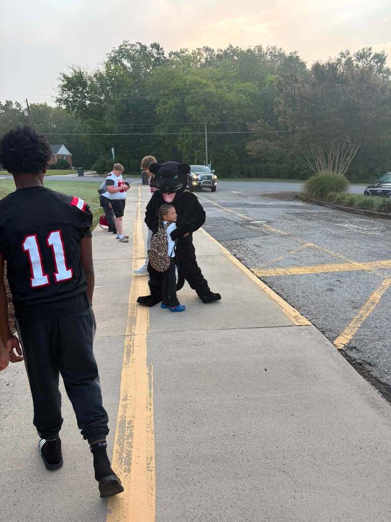 A community event takes place on a parking lot or street. In the foreground, a person wearing a black sports jersey numbered "11" stands on the left side of the pavement. In the center, an oversized black panther or big cat mascot costume interacts with a young child wearing a leopard-print outfit. Additional children and adults are visible along the sidewalk in the background. Parked vehicles are visible in the parking lot area behind the group. The setting is bordered by dense green trees and residential structures under an overcast sky. Yellow parking lot line markings are visible on the pavement.