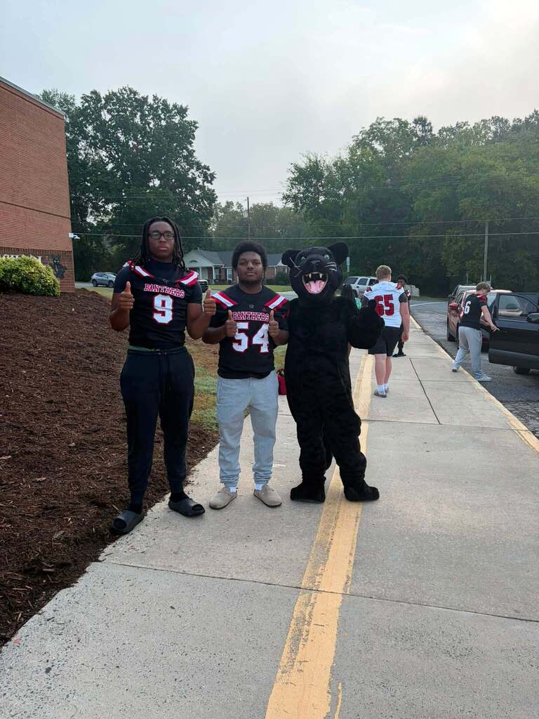 An outdoor photograph at a school athletic event shows three people in the foreground on a sidewalk: two individuals wearing black Panthers jerseys numbered 9 and 54 giving thumbs up, and one person wearing a black Panthers mascot costume with an animal head mask. Behind them are additional people in sports attire, a brick building, parked cars, a chain-link fence, trees, utility poles, and a partly cloudy sky, creating a festive school spirit atmosphere.
