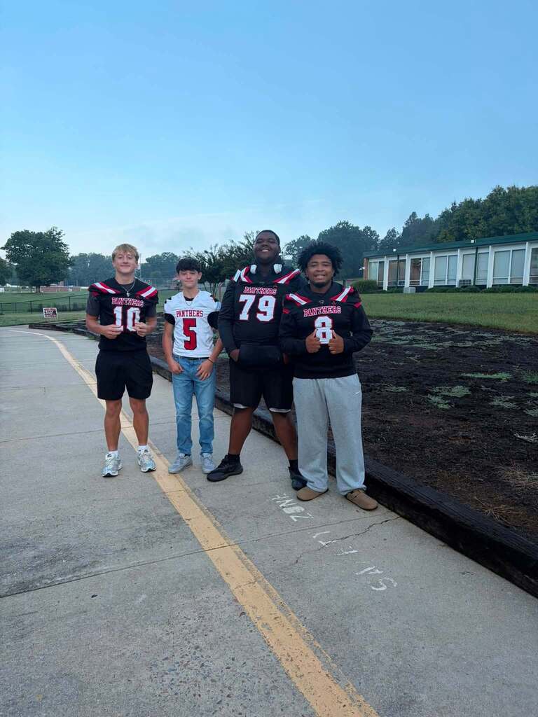 Four young people stand together on a concrete sidewalk outdoors. Three wear black Panthers football jerseys with red and white trim numbered 10, 79, and 8, while one wears a white Panthers jersey numbered 5 with blue jeans. They pose in front of a large white institutional building with green trees visible in the background. A grass field and clear blue sky are visible. A yellow center line runs along the sidewalk, with chalk writing visible on the pavement near their feet.