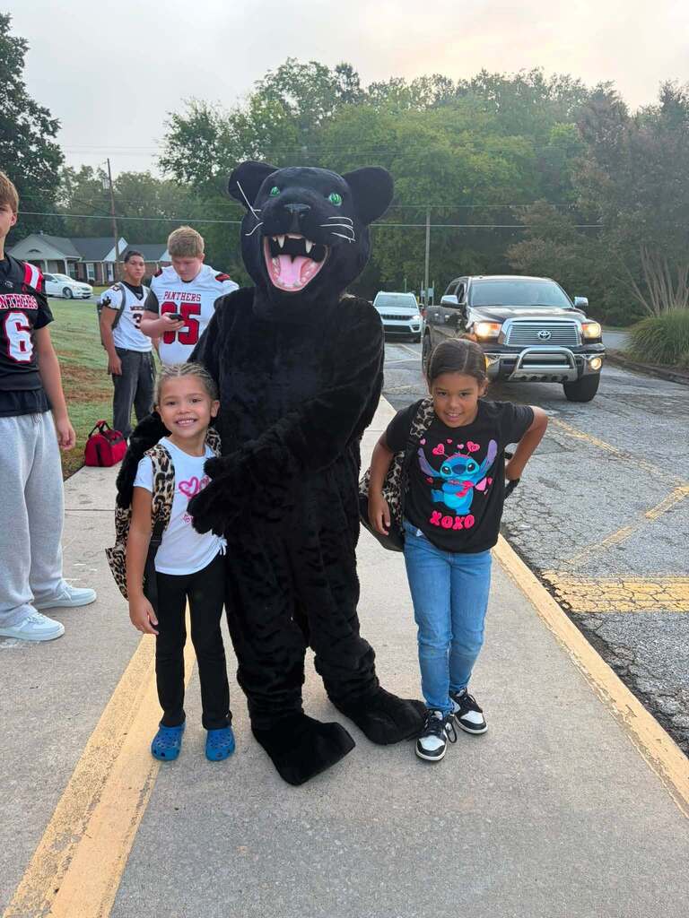 A large black panther mascot character with bright green eyes, white fangs, and an exaggerated happy expression poses with two young children on a paved road during an evening community event. In the background, other children and adults are visible, several wearing sports uniforms with "PANTHERS" printed on them. The setting includes vehicles, residential buildings, and trees, with lighting suggesting early evening hours. The casual, family-friendly atmosphere indicates a pep rally or pre-game gathering celebrating the local school sports program, with the Panthers team mascot interacting with community members.