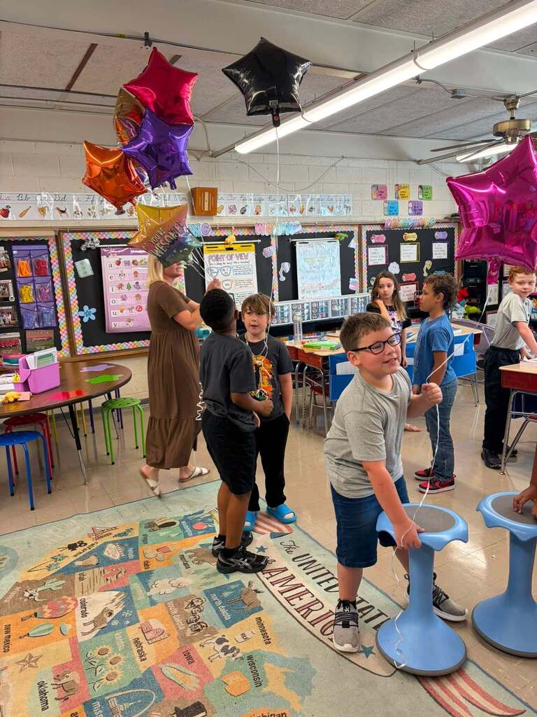 An elementary classroom hosts a festive celebration or special activity with multiple children and an adult instructor gathered together. Colorful foil star-shaped balloons in red, pink, purple, orange, yellow, and black hang from the ceiling, creating a celebratory atmosphere. Educational bulletin boards displaying student work, posters, and an alphabet banner decorate the walls. A large educational floor mat featuring instructional content is visible on the floor. Children use blue balance bikes or wobble seats as part of the activity. Colorful chairs in green and other colors and tables are arranged around the classroom perimeter. The adult instructor, wearing a brown dress, holds multiple balloons. The organized, festive setting supports group learning activities and student engagement.