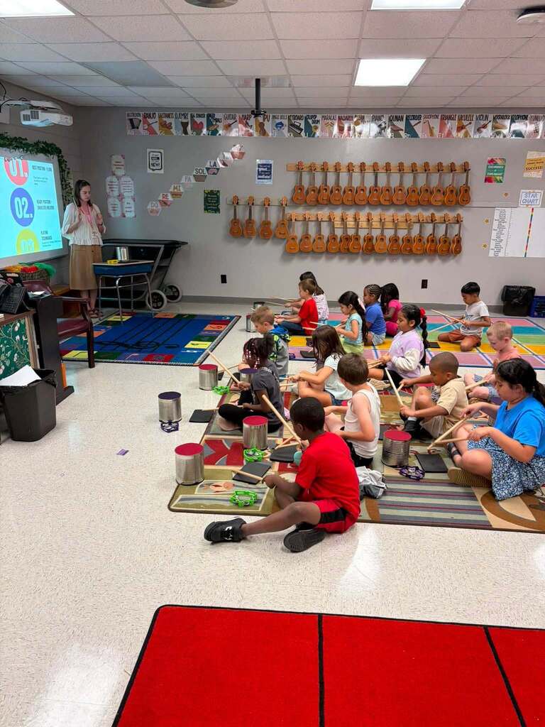 Approximately 20 elementary school children participate in an interactive music class, seated on colorful floor mats throughout the room. Each child holds a wooden drumstick and plays a metal percussion instrument, likely a steel drum or similar educational instrument, as part of a rhythm and percussion lesson. The well-equipped music classroom features over 50 acoustic guitars displayed on the wall in three horizontal rows, a teacher standing near a projector on the left, a wall-mounted projector screen displaying instructional content, colorful educational posters and materials, a decorative banner across the top of the wall, a multi-colored carpet mat in the center, metal trash bins, and wooden bookshelves with classroom supplies. The hands-on format and extensive instrument collection demonstrate a comprehensive elementary music education program.