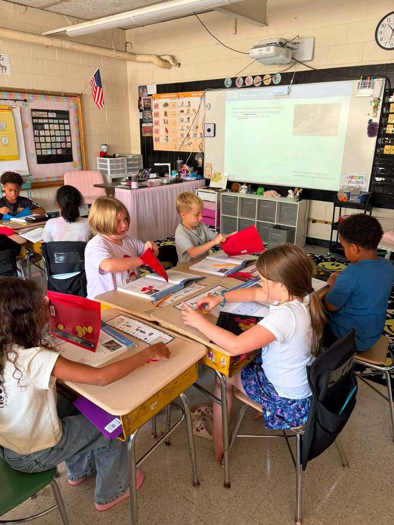 Elementary school students engage in a hands-on learning activity at their desks, working with red paper materials as part of a classroom project. The well-organized classroom features a teacher's desk with a pink table skirt, a smartboard displaying educational content, an American flag, colorful bulletin boards, educational displays, and wall-mounted storage with classroom materials. Ceiling-mounted fluorescent lighting, a projector, a wall clock, and multiple student desks create a bright, stimulating learning environment equipped with both traditional and digital learning tools for elementary-level instruction.
