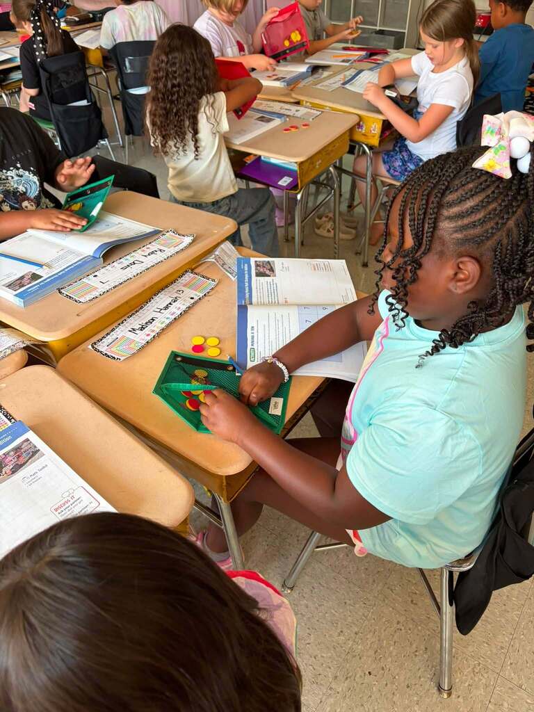 Elementary school students engage in active, hands-on learning at individual desks throughout the classroom. In the foreground, a student in a light blue shirt works with a green learning manipulative or game board containing colored counters with yellow and red pieces. Other students are seated at wooden desks with attached chairs or standing while working on various activities. Worksheets, printed materials, and educational resources are visible on desk surfaces. The scene captures an engaging learning environment with students working independently on different educational activities, with classroom furnishings and educational displays visible in the background.
