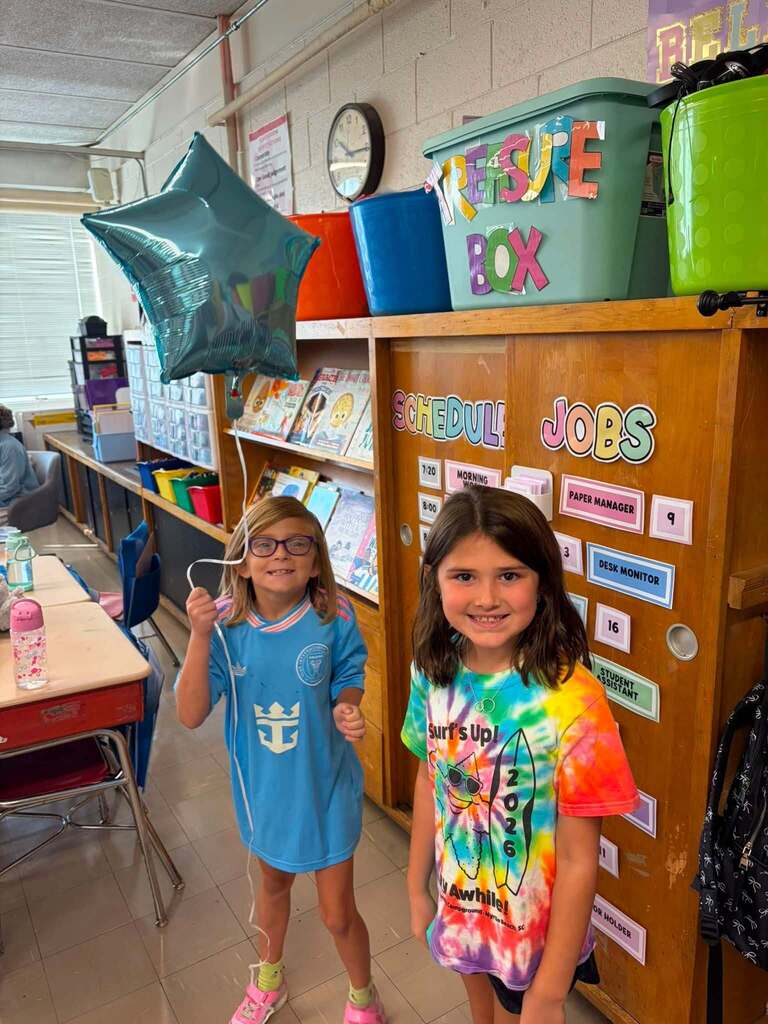 Two elementary school students stand in front of a wooden classroom "JOBS" cabinet displaying classroom helper assignments. The student on the left wears a blue shirt and holds a teal star-shaped balloon. The student on the right wears a colorful tie-dye shirt with "Surf's Up!" text and a sun graphic. Behind them, the cabinet displays visible job assignments including "MORNING HELPER," "PAPER MANAGER," "DESK MONITOR," and "STUDENT HELPER." A green "REUSE BOX" bin and colorful storage containers sit above the cabinet. Shelving below holds organized books, supplies, and classroom materials. The beige cinder block walls, fluorescent ceiling panels, wall clock, and organizational systems demonstrate typical elementary classroom management and job rotation systems.