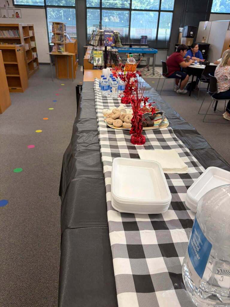 Library refreshment table set up for school event with black and checkered tablecloth. Table displays bottled water, juice, pastries, plates, and red centerpieces. Bookshelves and seated attendees visible in background, with colored floor markers on carpet.