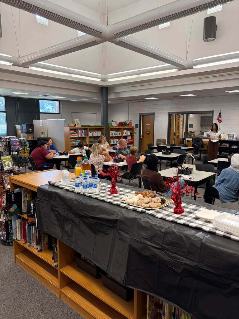 School library gathering with families seated at tables while a presenter stands nearby. Refreshment table in foreground displays bottled drinks, cookies, and pastries on black tablecloth with checkered runner. Bookshelves and American flag visible in background.
