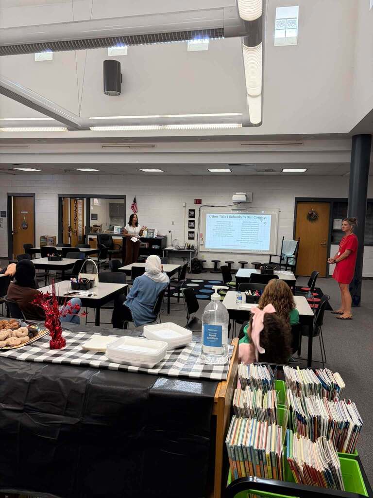 School library presentation showing projected slide titled 'Other Title I Schools in Our County' with list of school names. Adults and children seated at tables facing presenters at front of room. Refreshment table, bookshelves, and American flag visible in space.
