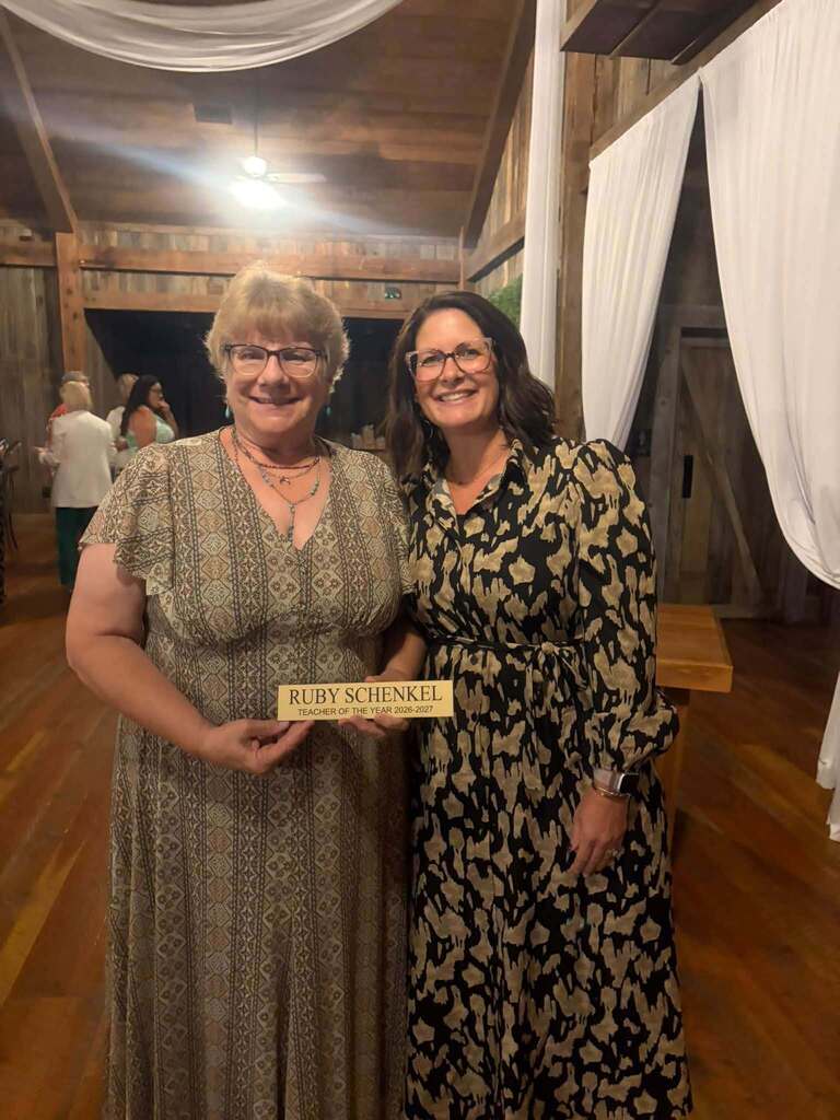 Two smiling women pose together indoors at a rustic wooden venue for a Teacher of the Year celebration. The woman on the left holds a small award sign reading "RUBY SCHENKEL TEACHER OF THE YEAR 2026–2027." White drapes, wooden walls, ceiling lights, and other attendees are visible in the background, creating a warm, celebratory atmosphere for this educational recognition event.