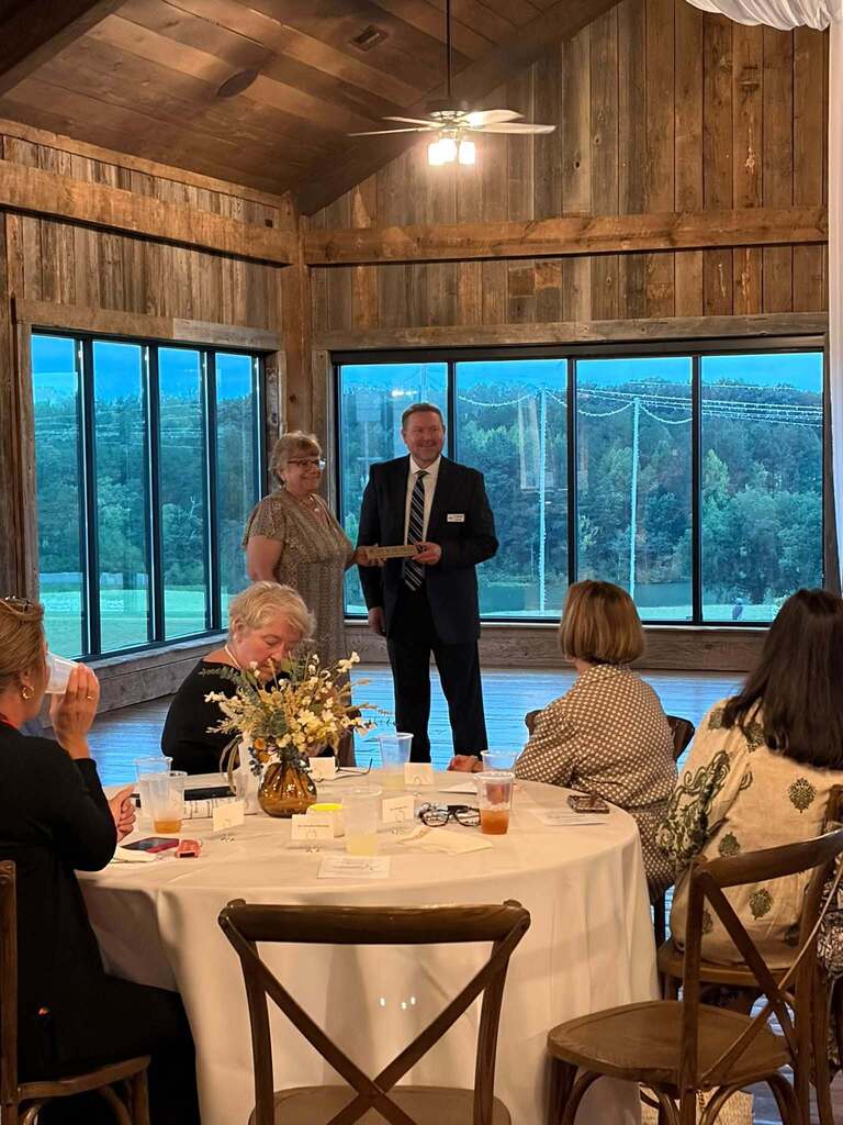 A formal indoor award ceremony is held in a rustic, wood-paneled venue with large windows overlooking a wooded landscape and water. A man in a suit stands at the front holding a small plaque or award while addressing the gathering. Several seated attendees watch from a round table decorated with flowers, place settings, drinks, and name cards. The formal presentation setting and elegant table arrangements indicate a special recognition or appreciation event.