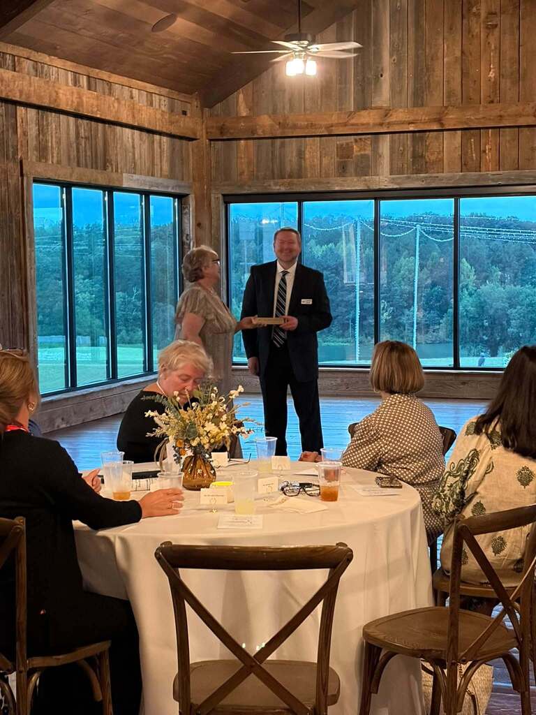 A formal indoor award ceremony takes place in a rustic wood-paneled venue with large windows overlooking a wooded landscape and a swimming pool or open lawn. A man in a dark suit and tie stands beside a woman, appearing to present or receive an award or recognition item. Several seated guests gather around a round table covered with a white tablecloth and decorated with flowers, drinks, place cards, and personal items. The elegant setting and formal attire suggest a special recognition event or celebration.