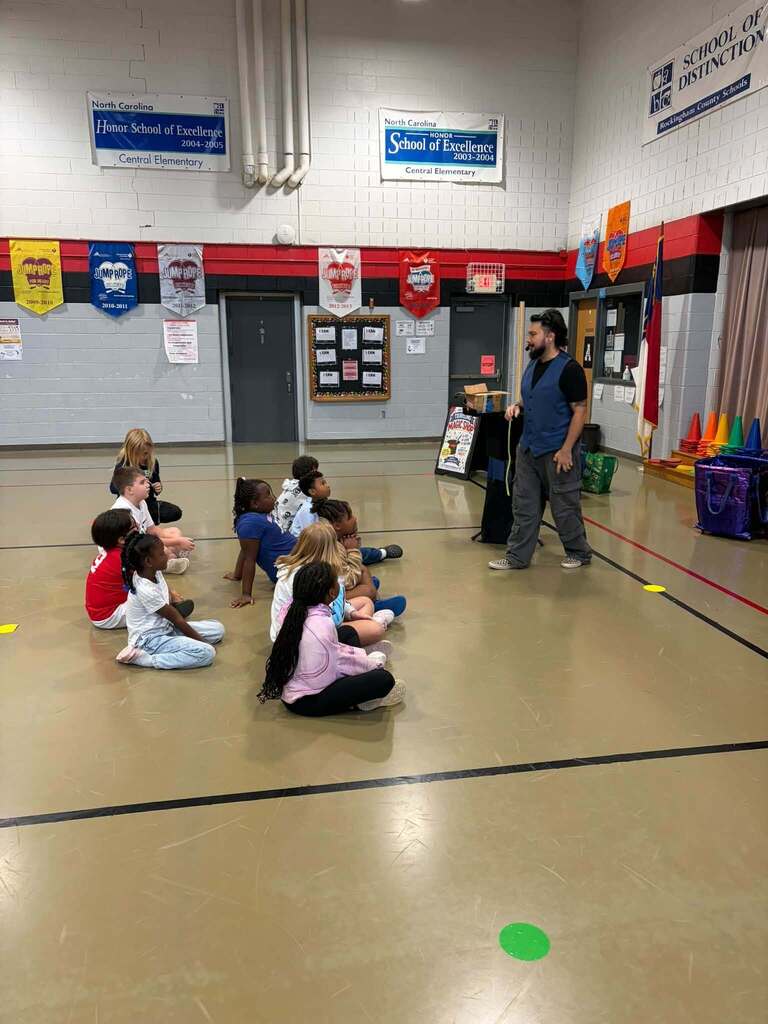 A group of elementary school children sit on a gym floor facing an adult instructor during what appears to be a magic show presentation. The gymnasium walls display multiple award plaques and colorful banners recognizing Central Elementary as a "North Carolina Honor School of Excellence" for 2003–2004 and 2004–2005, along with a "School of Distinction" designation and "Jump Rope for Heart" recognition. Cones and other activity equipment are visible in the gym, indicating an active learning environment focused on physical fitness and wellness education.
