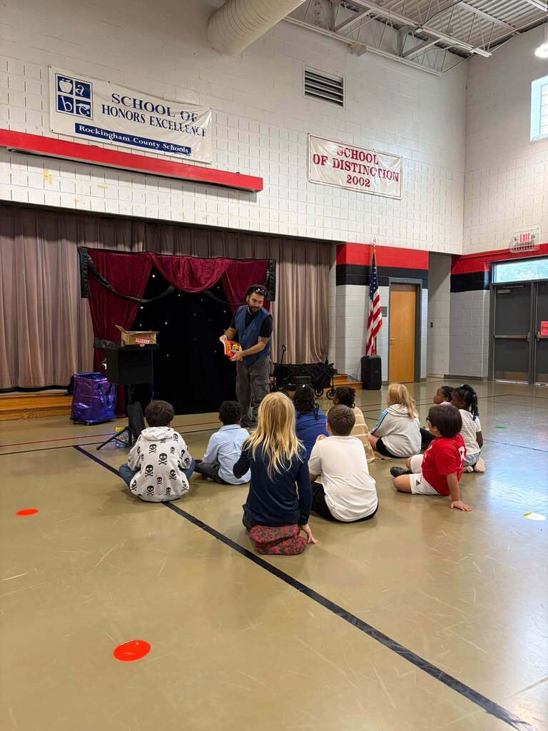  A man demonstrates or presents something to a group of children seated on the floor in a school gym or auditorium during an educational assembly. Behind him is a small curtained stage with a table and equipment. An American flag and school doors are visible in the background. Wall banners display "SCHOOL OF HONORS EXCELLENCE Rockingham County Schools" and "SCHOOL OF DISTINCTION 2002," highlighting the school's academic recognition and excellence. The formal presentation setting indicates an organized educational or motivational program for students.