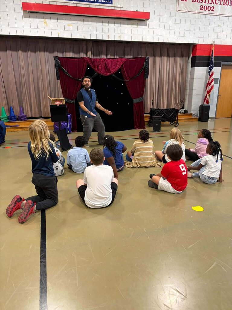 A man stands in front of a curtained, theatrical-looking stage area while a group of children sit on the gym floor facing him during what appears to be a magic show assembly. An American flag, speaker, and equipment cart are positioned nearby. Partially visible text on the wall reads "County Schools" and "OF DISTINCTION 2002," indicating school recognition and achievement. The setting suggests a formal presentation or instructional session in the school gymnasium.