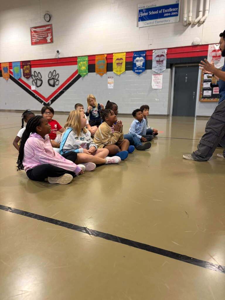 Several elementary school children sit on a gym floor looking toward an adult instructor standing on the right, who gestures as if speaking to the group during a magic show presentation. The gym wall behind them features colorful pennants, paw-print decorations representing the school mascot, and a visible sign reading "North Carolina Honor School of Excellence 2004–2005 Central Elementary," showcasing the school's academic achievements and school spirit.