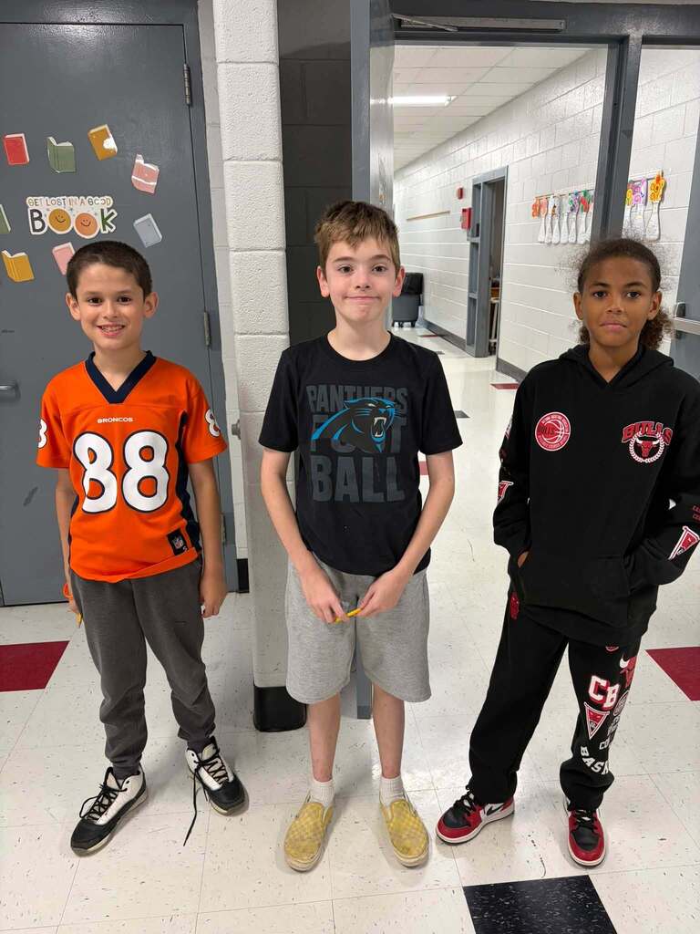 Three boys stand together in a school hallway wearing sports-themed clothing representing different teams. The left student wears a "BRONCOS" jersey numbered 88, the center student wears a "PANTHERS FOOTBALL" shirt, and the right student wears a "BULLS" hoodie with Chicago Bulls logos. Behind them, a classroom door is decorated with book stickers and displays the message "GET LOST IN A GOOD Book," promoting literacy and reading engagement. The image captures students expressing their individual sports team preferences while in a school environment that encourages reading and learning.