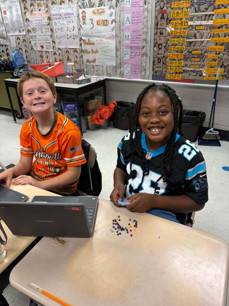 Two smiling students sit together at a classroom table during a learning activity. One student wears an orange baseball jersey while the other wears a black-and-blue Carolina Panthers jersey, demonstrating school spirit and team pride. A Lenovo laptop and small colorful learning manipulatives are visible on the table. Classroom posters and a schedule are displayed in the background, creating an organized educational environment that supports both technology-integrated learning and student collaboration.