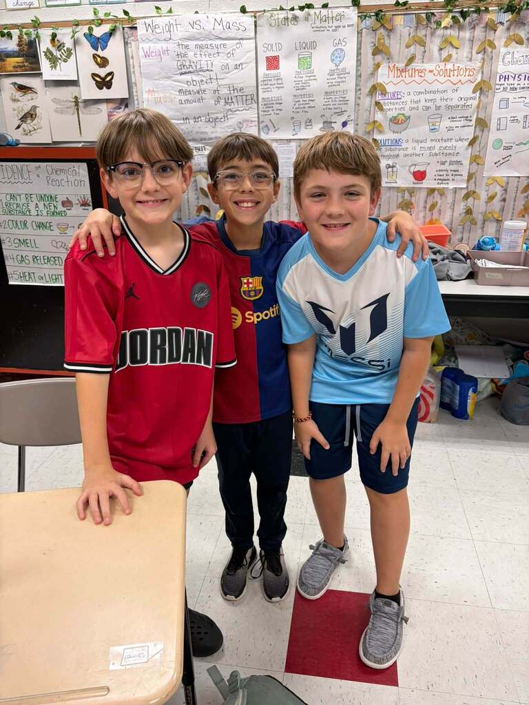Three school-age boys stand together in a science classroom, smiling with their arms around one another while wearing sports jerseys. 