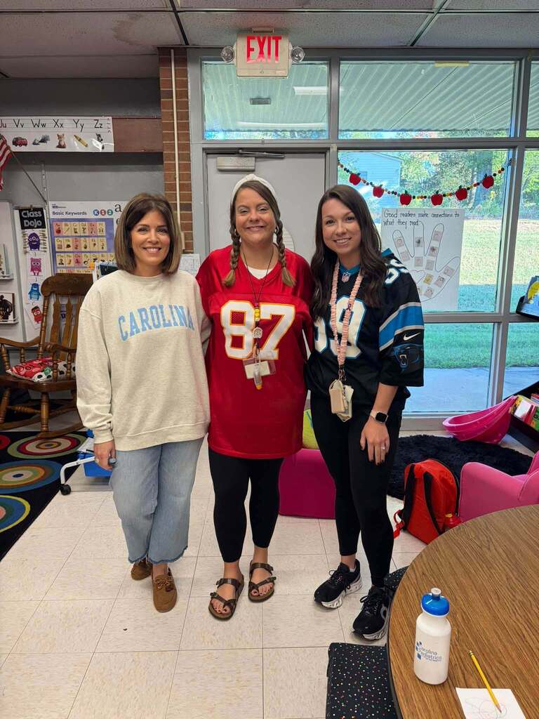 Three smiling women stand together in an elementary classroom showing school spirit by wearing sports apparel. One woman wears a red jersey numbered 87, while the others wear Carolina and Carolina Panthers clothing. The classroom features educational posters including one reading "Good readers make connections to what they read," along with "ClassDojo" and "Basic Keywords" signage. Colorful rugs, a table, children's seating, and an EXIT sign are visible in the background. The word "CAROLINA" is prominently displayed, demonstrating staff camaraderie and team spirit within the learning environment.