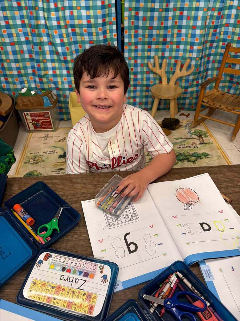 A smiling child sits at a classroom table wearing a white pinstriped baseball shirt, engaged in hands-on learning activities. The table is covered with open activity books, crayons, markers, scissors, and pencil cases. One workbook page displays a colored drawing of an apple, while another shows a large numeral 6 with practice drawings. A colorful checkered curtain and child-sized furniture are visible in the background, creating a bright, welcoming early childhood classroom environment that supports creative and educational development.