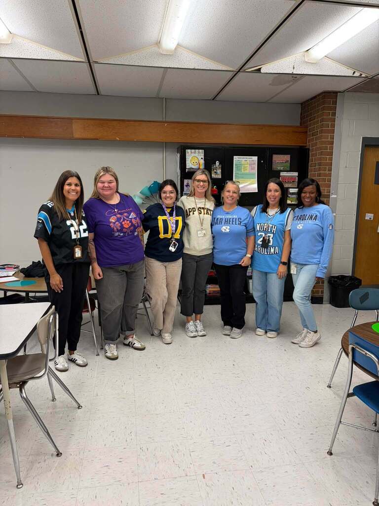Seven women stand together smiling in an elementary classroom, displaying school and college pride through their apparel. Several wear college-themed clothing featuring "APP STATE," "TAR HEELS," "NORTH CAROLINA," and "CAROLINA" text, celebrating their alma maters and creating a sense of community. The classroom setting includes student desks and chairs, a bulletin board, and a drop ceiling, with the staff members' casual, spirited attire demonstrating camaraderie and a welcoming school culture.