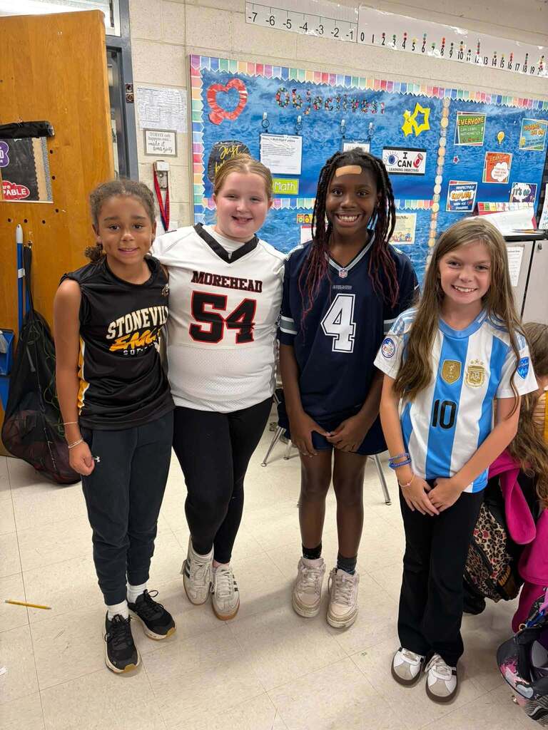 Four school-age girls stand together in a classroom smiling while wearing various sports jerseys, demonstrating school spirit and team pride. The jerseys include "STONEVILLE," "MOREHEAD" with the number 54, and an Argentina-style blue-and-white jersey with the number 10. The classroom background features a colorful bulletin board and a number line, creating an engaging elementary learning environment where students express their individual interests and team affiliations while participating in their education.