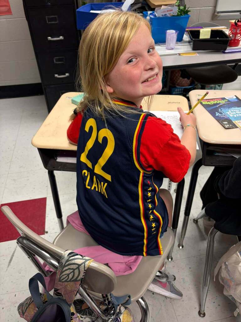 A smiling student with blonde hair sits at a school desk in a classroom wearing a red shirt and a navy jersey with yellow and red trim displaying the number 22 and the name "CLARK." The student holds a pencil and has paper on the desk, engaged in classwork. Other desks, classroom supplies, and a backpack are visible nearby, capturing a typical learning moment in an elementary classroom where students show school spirit through sports apparel while completing their academic work.