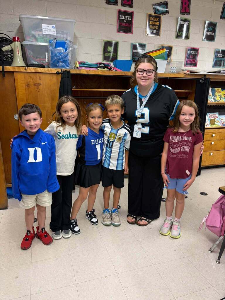 An adult and five children stand together in a classroom, with several wearing sports-themed clothing. Behind them, a prominent classroom poster displays positive character education messages reading: "BELIEVE IN YOURSELF," "WORK HARD HAVE FUN," "BE HONEST," "RESPECT OTHERS," "ALWAYS BE KIND," "ASK," "POSITIVE," and "TRY A LITTLE KINDNESS." Shelves and classroom materials are visible in the background. The combination of sports spirit and character education messaging demonstrates the school's commitment to both academic excellence and social-emotional learning, creating a supportive classroom culture that values kindness, respect, and personal growth.