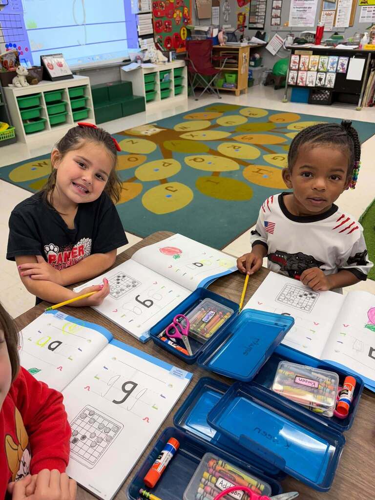Three young children sit at a classroom table engaged in early math learning activities. Open workbooks displaying number-writing exercises and counting grids are spread on the table along with pencils, crayons, scissors, and glue. The children work on their activities while seated near a colorful educational rug featuring large numbered shapes in the background, creating an engaging early childhood learning environment focused on numeracy skills development.