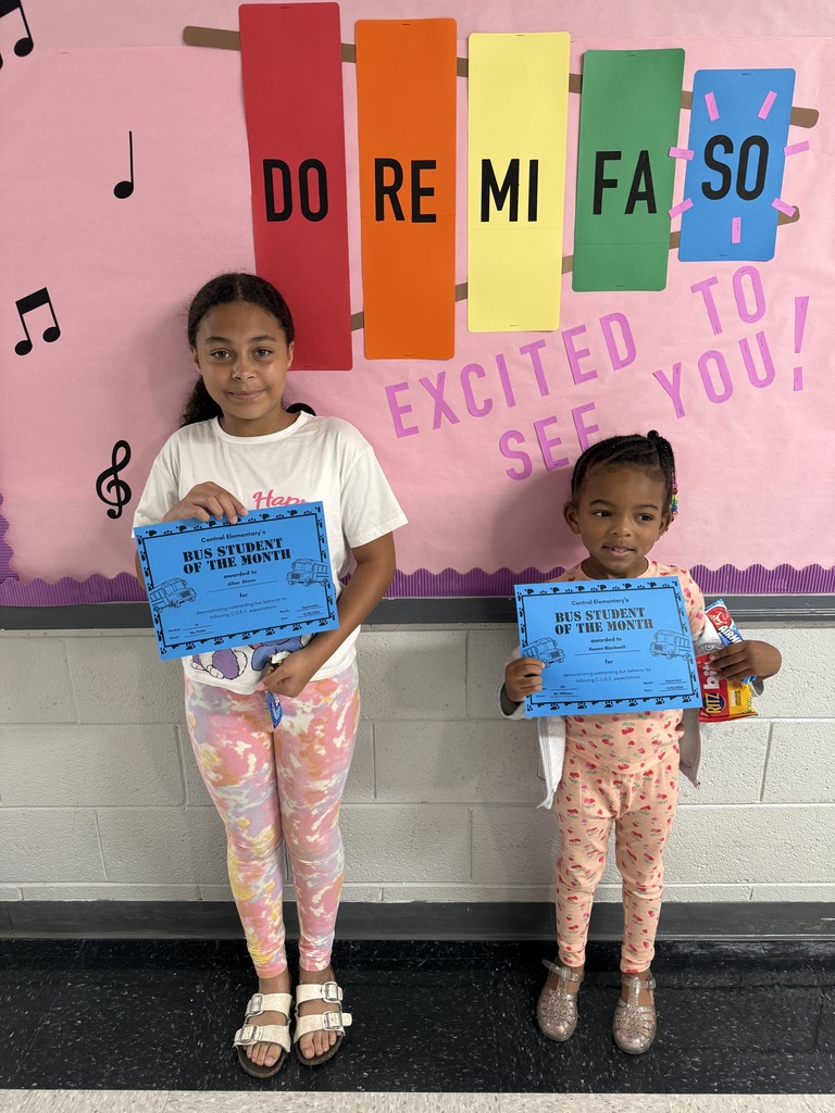 Two children stand proudly in a school hallway holding blue certificates recognizing them as "Central Elementary's BUS STUDENT OF THE MONTH." Behind them, a vibrant pink classroom bulletin board is decorated with colorful musical notes and displays the welcoming messages "DO RE MI FA SO" and "EXCITED TO SEE YOU!" The image celebrates positive student behavior and recognition for exemplary conduct on school transportation, while the musical-themed classroom display creates a warm, inviting learning environment. This recognition program highlights the school's commitment to acknowledging students who demonstrate respect, responsibility, and good citizenship during their daily bus rides.