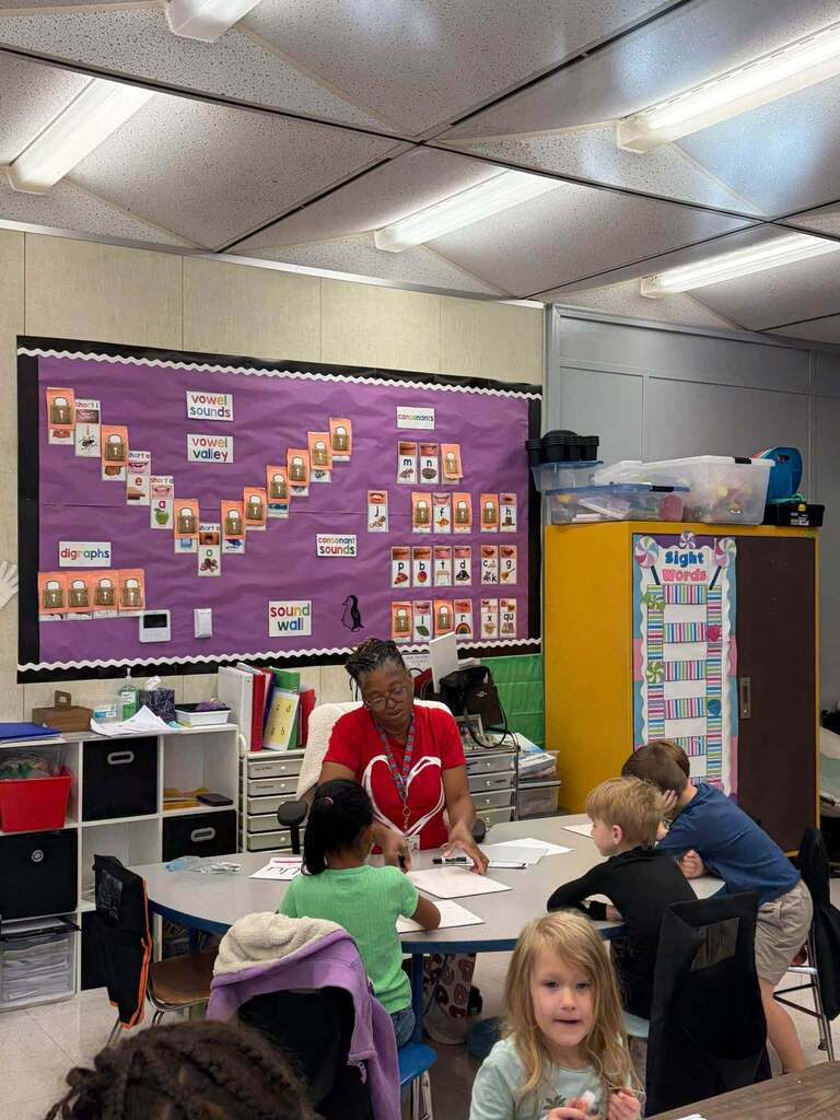 A teacher works with several children seated around a table during small-group literacy instruction. Behind them, a purple classroom display features a comprehensive phonics "sound wall" with organized sections labeled "vowel sounds," "vowel valley," "consonant sounds," and "digraphs," along with letter-sound cards. A nearby cabinet displays a "Sight Words" section with storage bins on top. The structured phonics instruction environment demonstrates the school's commitment to evidence-based literacy instruction using systematic approaches to reading development, with visual supports that help students connect letters to sounds.
