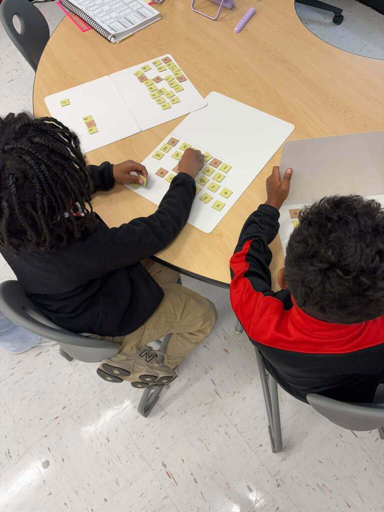 Two students sit at a table engaged in hands-on phonics learning, arranging small letter tiles on white boards. The tiles display individual letters and letter combinations including "a," "b," "c," "d," "f," "h," "i," "k," "m," "n," "o," "r," "s," "t," "u," "v," "y," "z," and "qu." A notebook and classroom supplies are also visible on the table. The manipulative-based learning activity allows students to physically build and manipulate words, supporting their understanding of letter-sound relationships and word construction through tactile, interactive literacy instruction.